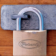Primo piano di un lucchetto in ottone Potent agganciato a una chiusura in metallo zincato su una porta in legno. Il logo Potent è visibile al centro del corpo del lucchetto