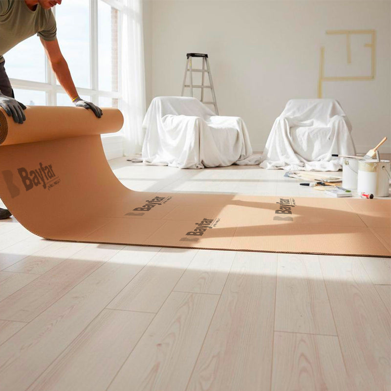 Uomo che srotola un lungo telo protettivo in cartone ondulato Bayfar su un pavimento di un ambiente da dipingere con mobili coperti e attrezzatura da pittura